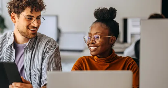 A man and a woman are sitting at a desk with open laptops in from of them. They are looking at one another and smiling.