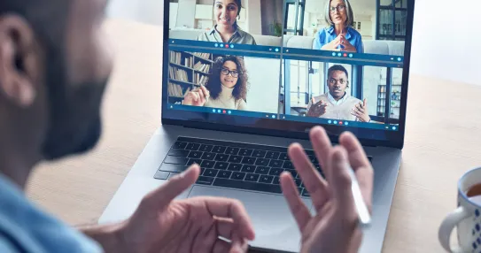 A man looks at a laptop on his desk. The laptop shows he is in a video call with many other people.