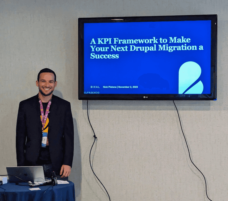 Bixaler Nick Pistone, a smiling man with dark hair, stands behind a table with his laptop while behind him a television projects the title of his Drupal GovCon presentation, “A KPI Framework to Make Your Next Drupal Migration a Success.