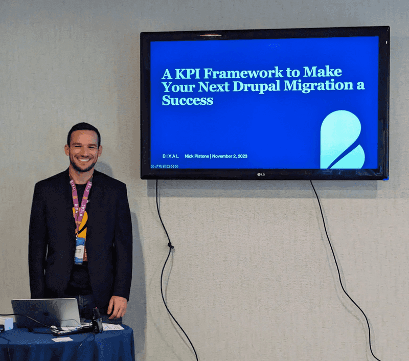 Bixaler Nick Pistone, a smiling man with dark hair, stands behind a table with his laptop while behind him a television projects the title of his Drupal GovCon presentation, “A KPI Framework to Make Your Next Drupal Migration a Success.