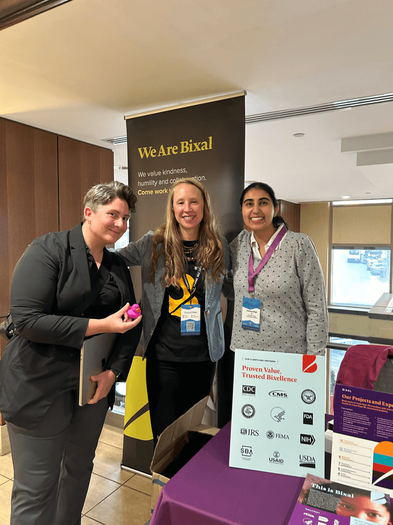 Three Bixal team members pose for a photo by Bixal’s colorful booth at the Drupal GovCon conference. One holds up a purple rubber duck.