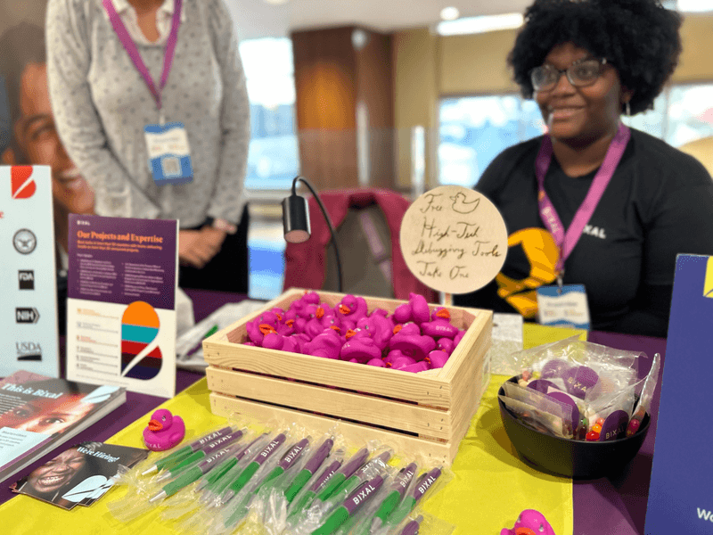 Bixaler Felicia Odie sits behind the Bixal table at Drupal GovCon. On the table are Bixal-branded swag items for conference goers, including purple rubber ducks, pens and colorful jelly beans.